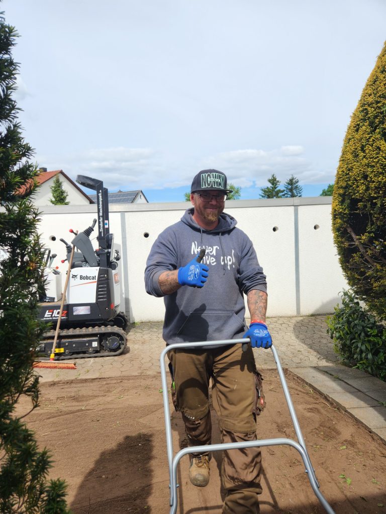 Ein Foto vom Inhaber mit einer Gartenwalze beim vorbereiten für die verlegung eines Rollrasens im Hintergrund ein Minibagger.